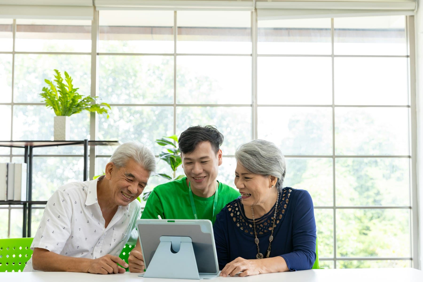 A young caregiver helping an older couple with a tablet at a table by the window