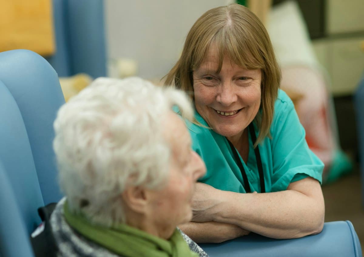 A caregiver in green scrubs smiling beside an elderly woman in an armchair