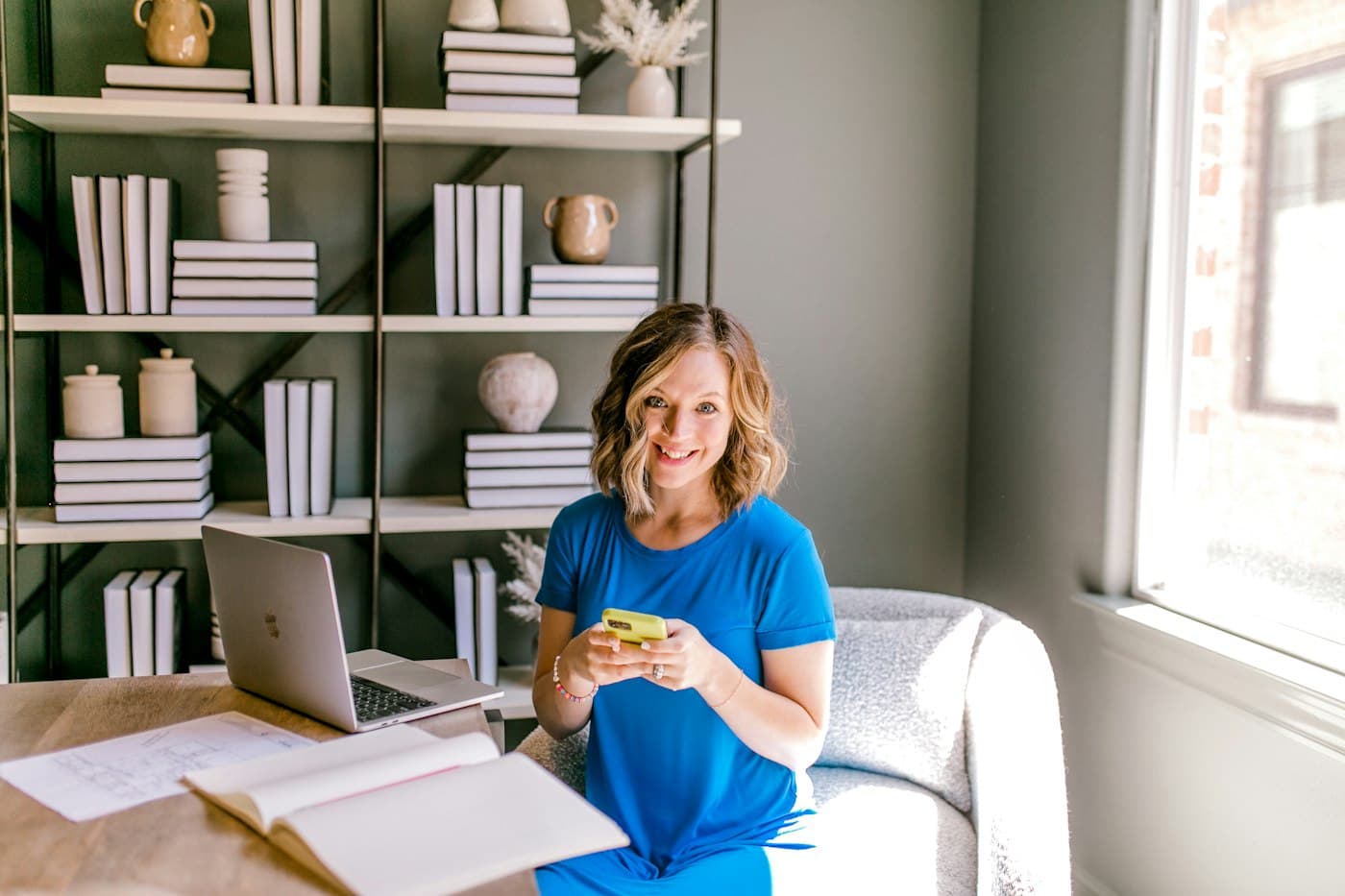 A woman smiling at her phone at a desk with an open laptop